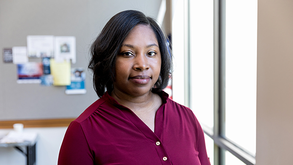 A person wearing a burgundy blouse stands in a well-lit room with a bulletin board in the background. They have shoulder-length dark hair and are looking directly at the camera.