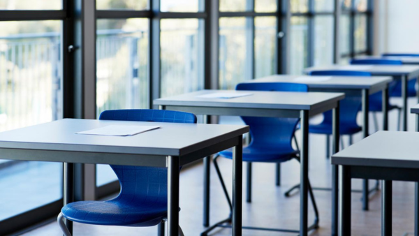 A brightly lit classroom with several empty blue chairs and desks. Each desk has a piece of paper on it. Large windows line one side of the room, allowing natural light to illuminate the space.