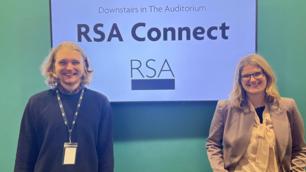 Two people stand in front of a teal wall with a large screen displaying "Downstairs in The Auditorium RSA Connect" and the RSA logo. Both are smiling, the person on the left has long hair, and the person on the right has glasses.