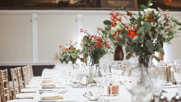 A long dining table is elegantly set for an event, adorned with tall floral centerpieces featuring red and white flowers. The table is lined with white chairs, glassware, and folded napkins in a softly lit room.