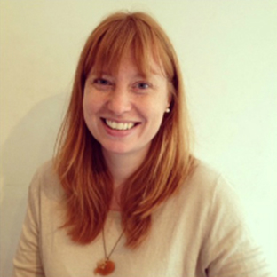 A person with long red hair smiles warmly at the camera, wearing a beige top and a necklace with a round pendant. The background is plain and light-colored.