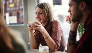 A woman, smiling, holds a coffee cup while seated at a table with a man. They are indoors, near a window with light streaming in. The atmosphere is relaxed and warm, with flowers visible in the background.