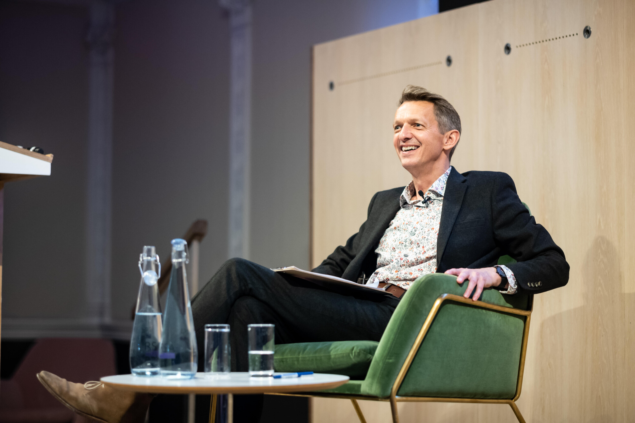 A person sitting in a green chair, smiling during a talk. They are holding a folder and wearing a dark suit with a patterned shirt. A table with a water carafe and glasses is on the left. The background features a wooden partition.