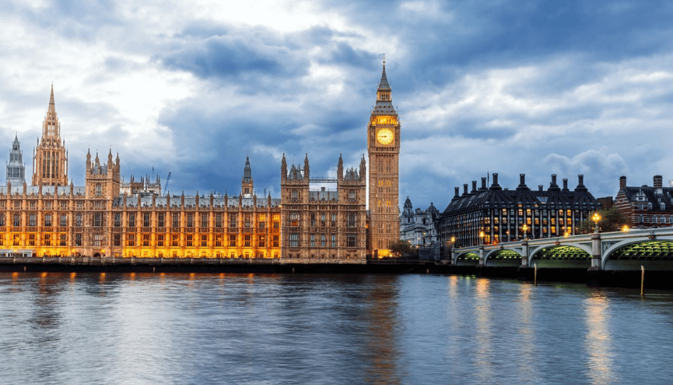 The image shows the Houses of Parliament and Big Ben in London at dusk. The buildings are illuminated with lights, reflecting off the River Thames. The sky is cloudy with a soft blue hue.
