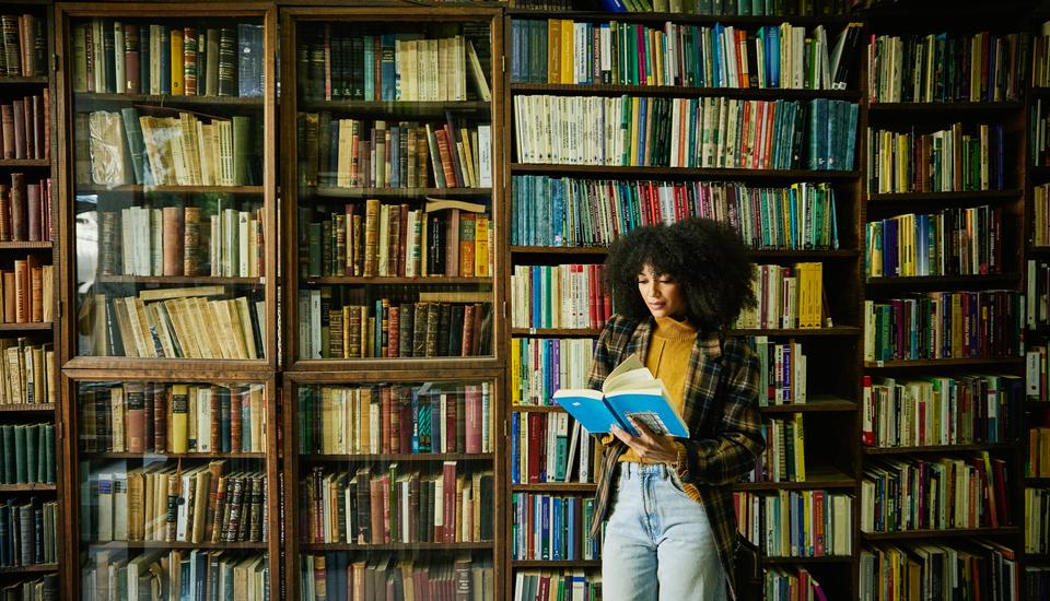 A woman with curly hair stands reading a book in front of tall, filled bookshelves. They wear a plaid coat, mustard sweater, and jeans. The setting appears to be a cozy library or bookstore.