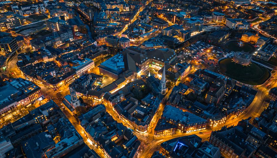 Aerial view of a city at night with illuminated streets forming a network of glowing lines. Buildings are densely packed, and a notable structure with bright lights stands out in the center. Streetlights and car headlights create a vibrant cityscape.