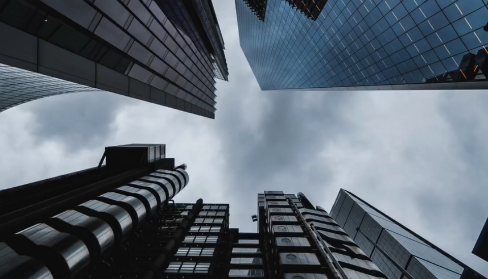 A low-angle view of modern skyscrapers with glass and steel facades, converging towards a cloudy sky. The buildings vary in design and height, creating a dynamic architectural composition against the overcast backdrop.
