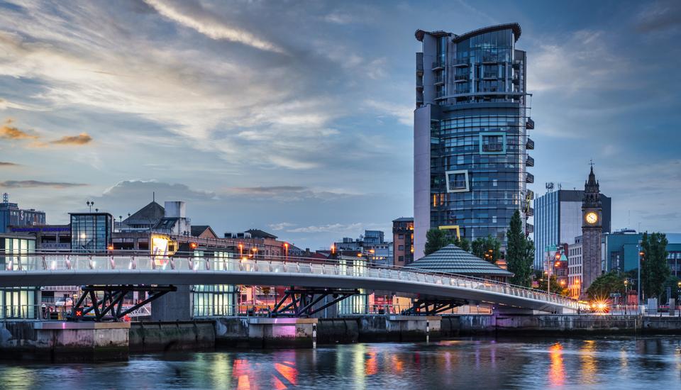 A modern cityscape at sunset features a curved pedestrian bridge over a river, leading to a tall, contemporary building. The sky is partly cloudy, and city lights reflect on the water, creating a serene urban atmosphere.