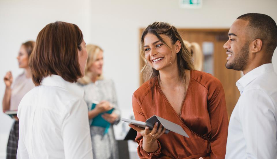 A group of people engaged in conversation in an office setting. A woman in a rust-colored blouse holds a tablet and smiles at another woman in white. In the background, others are talking and holding papers.