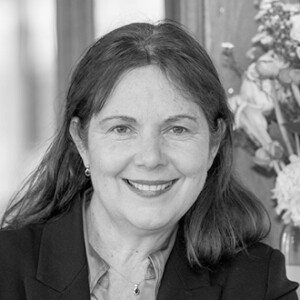 A woman with long dark hair and a blazer smiles at the camera. She is indoors, with a bouquet of flowers visible in the background. The image is in black and white.
