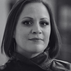Black and white portrait of a person with straight shoulder-length hair, wearing a jacket with the collar up. They have a neutral expression, and the background is softly blurred.