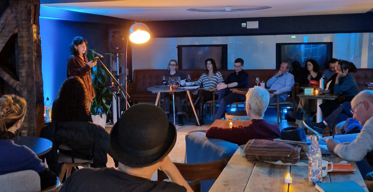 A person stands on stage performing in front of an audience in a cozy venue with dim lighting. People are seated at tables with drinks, and a hanging lamp illuminates the performer. Candles are lit on the tables, enhancing the relaxed atmosphere.