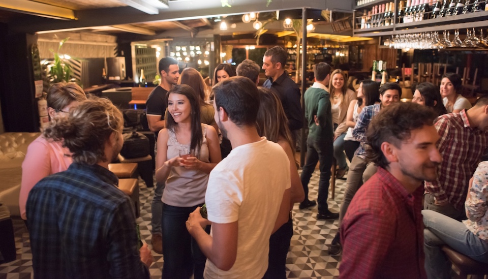 A lively bar scene with a group of people socialising. Men and women are engaged in conversation, holding drinks. The setting features a cozy ambiance with dim lighting, patterned floor tiles, and shelves of bottles behind the bar.