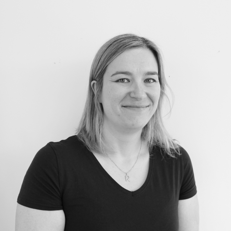 A person with shoulder-length hair, wearing a black t-shirt and necklace, smiles slightly while standing in front of a plain white background. The image is in black and white.