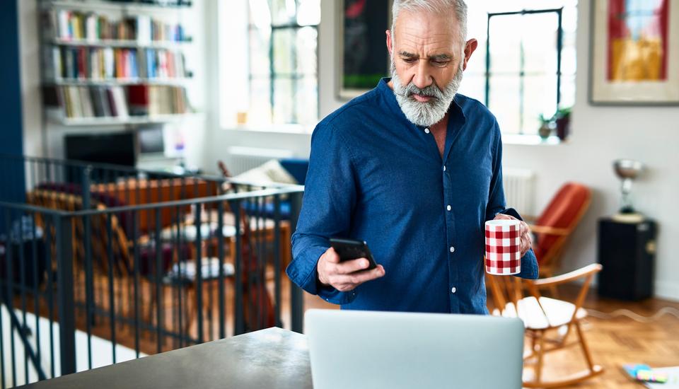 A bearded man in a blue shirt holds a red and white checkered mug while checking his phone. He stands in a modern, spacious room with a laptop on the table and a bookshelf in the background.