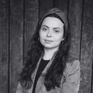 An image of Sophia Smith Galer with long dark hair, wearing a headband and hoop earrings, is posing in front of a wooden wall. They are dressed in a patterned jacket, conveying a confident and relaxed expression. The image is in black and white.