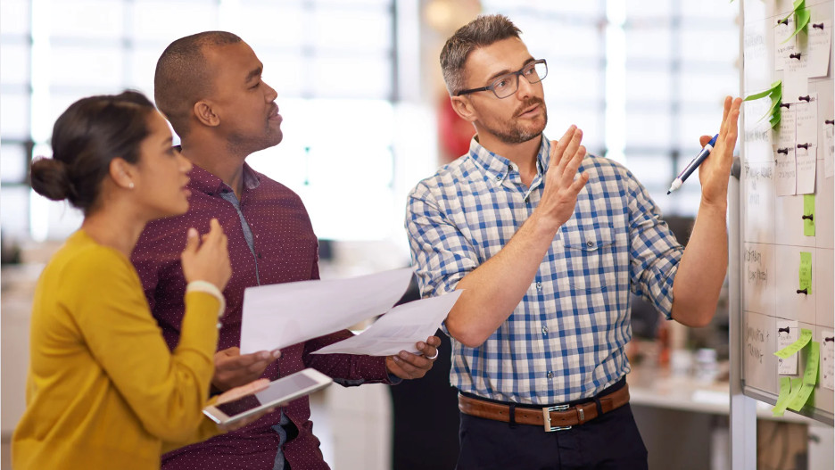 Three people are engaged in a discussion in an office. A man on the right gestures at a whiteboard with notes, while the other two listen attentively, holding papers and a tablet. They appear focused and collaborative.