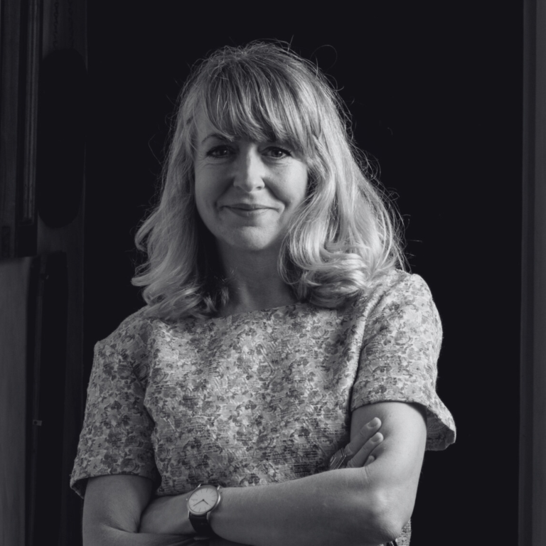 Black and white portrait of Alecky Blythe with shoulder-length hair, smiling and standing with arms crossed. She is wearing a patterned blouse and a watch on her left wrist, set against a dark background.