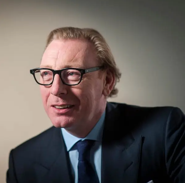 Benny Higgins with blonde hair and glasses, wearing a dark suit with a blue shirt and tie, is smiling and looking slightly to the side against a neutral background.