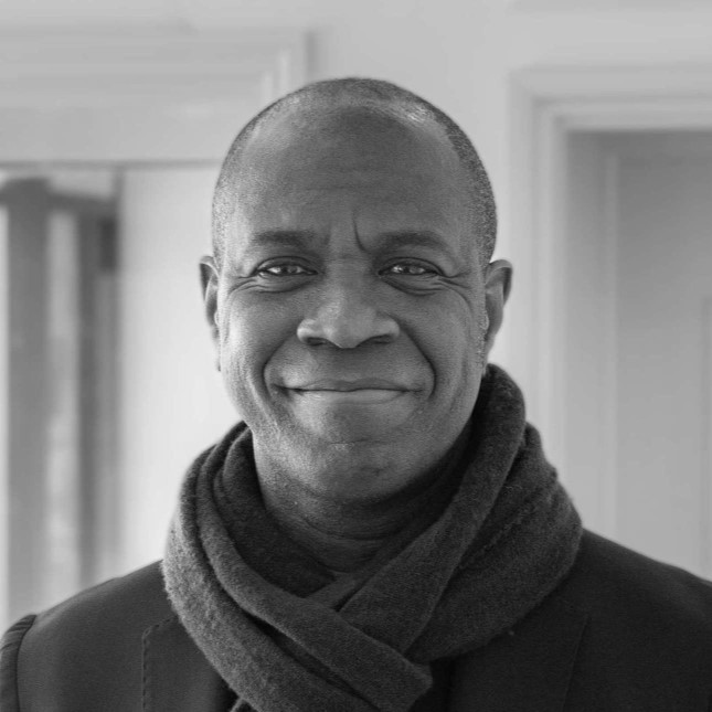 An image of Clive Myrie smiling, wearing a dark scarf and coat stands in front of a blurred indoor background. The grayscale image captures their friendly expression with a softly lit setting.