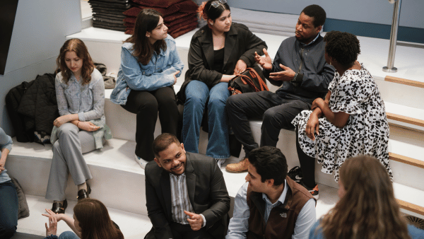 A group of people are sitting on steps, engaged in conversation. They are dressed in casual and business attire. Some are gesturing while talking, and others are listening attentively. A stack of folded clothes is visible in the background.