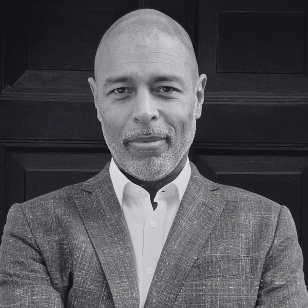 An image of Dawid with a bald head and beard is wearing a light-colored shirt and dark blazer. They are standing against a dark paneled backdrop, with arms folded and a neutral facial expression. (Black and white image).