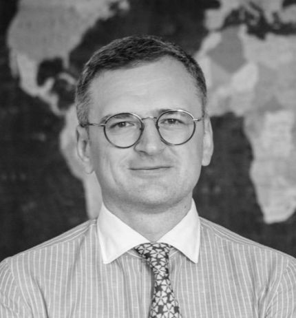 An image of a Dmytro Kuleba with short hair and glasses is wearing a striped shirt and patterned tie, standing in front of a blurred world map background. The image is in black and white.