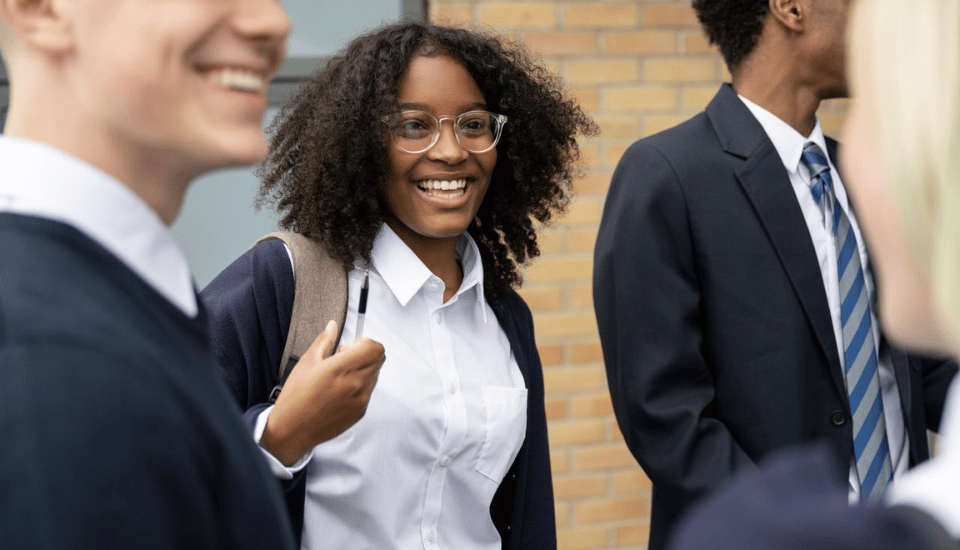 A group of students in school uniforms smiling and talking outdoors. One student in focus is wearing glasses and holding a backpack strap. A brick building is visible in the background.