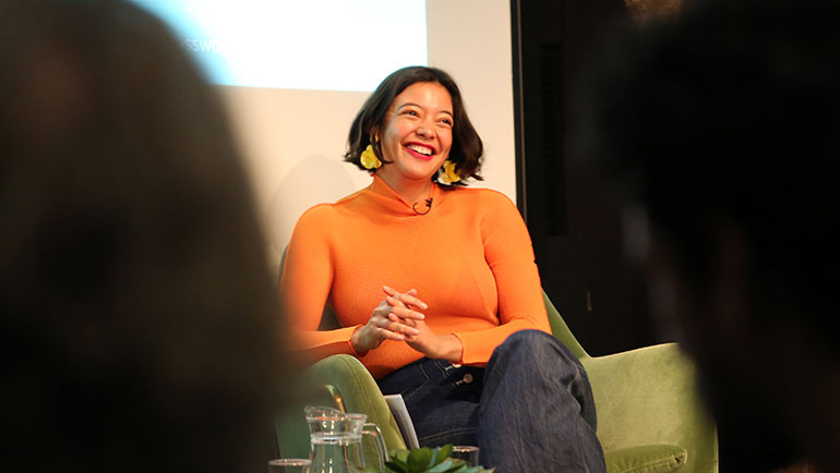 A person wearing a bright orange sweater and yellow earrings smiles while seated on a green chair. They are on a stage or panel, with a blurred audience in the foreground and a projection screen in the background.