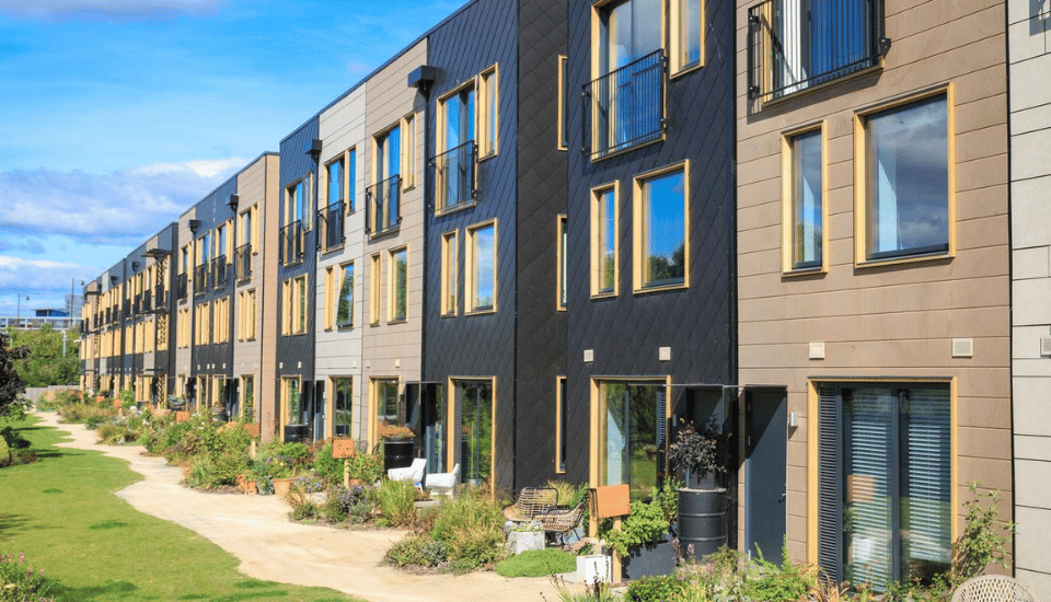 A row of modern townhouses with a mix of black and beige facades, large windows, and small front gardens. A pathway runs along the green lawn in front of the buildings, under a bright blue sky.