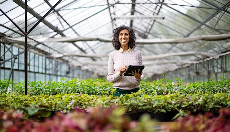 A person stands in a greenhouse surrounded by various plants, holding a tablet and smiling. The greenhouse structure is made of metal and glass, allowing natural light to flood the space. The person is wearing a light sweater.