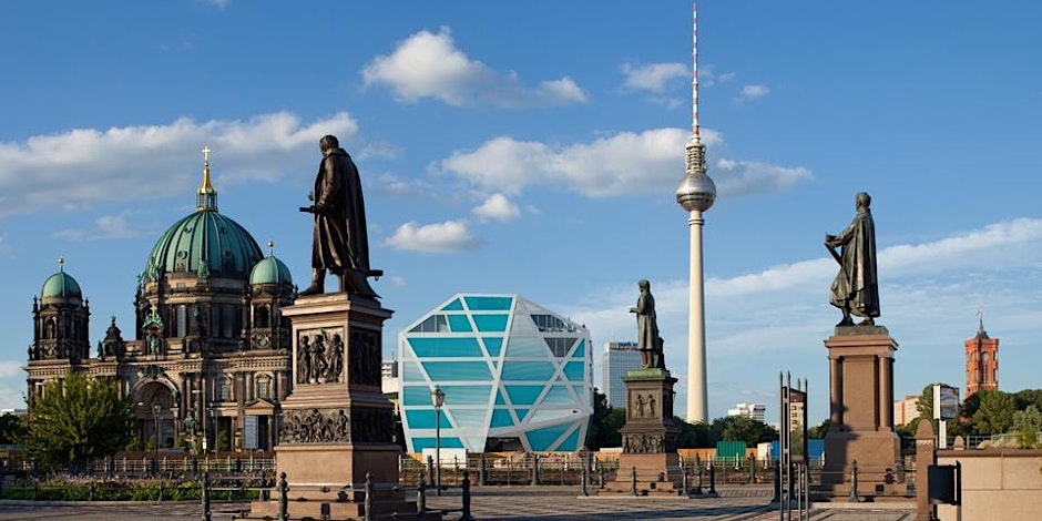 Statues stand in the foreground with a view of the Berlin Cathedral, a modern glass building, and the Fernsehturm tower in the background under a blue sky with scattered clouds.