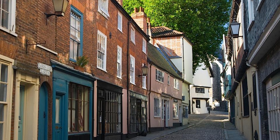 A quaint cobblestone street lined with colorful, historic buildings and rustic street lamps on a sunny day. A lush green tree is visible at the end of the street, with soft shadows suggesting a peaceful atmosphere.