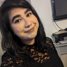 An image of Iqra Choudhry with shoulder-length dark hair and a subtle smile is wearing a black lace top. They are indoors, with a TV and some books or boxes visible in the background.