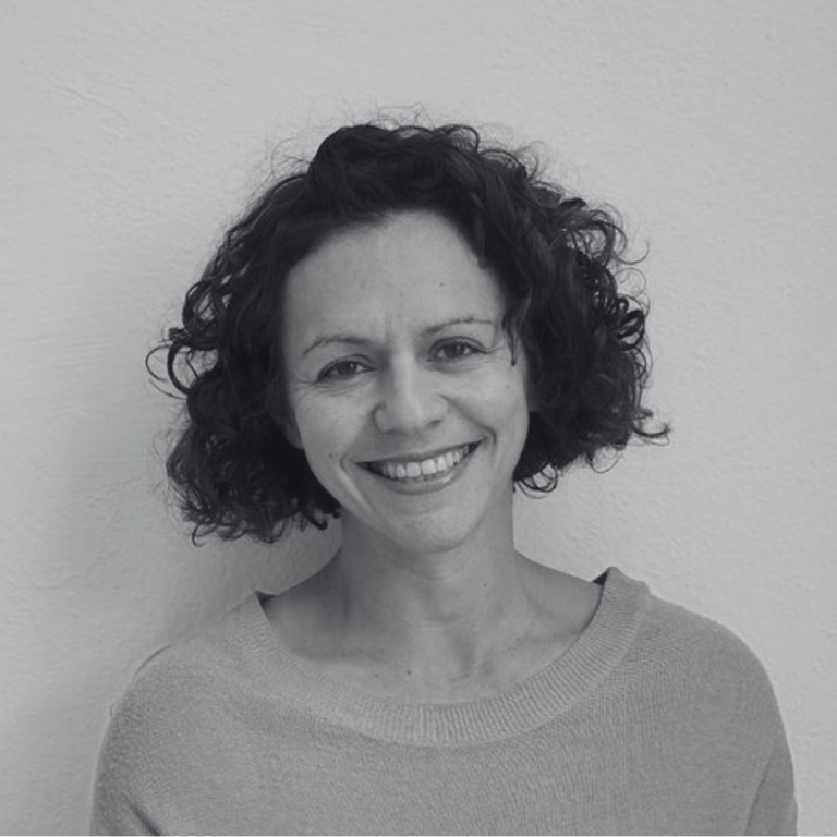 An image of Josephine Burton with curly hair smiles while facing the camera. They are wearing a light-colored top and standing against a plain background. The image is in black and white.