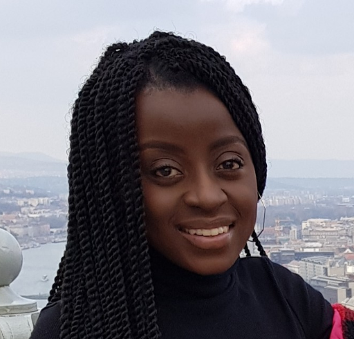 Lauryn Mwale with long braided hair, wearing a black top, smiles in an outdoor setting with a cityscape and river in the background under a cloudy sky.