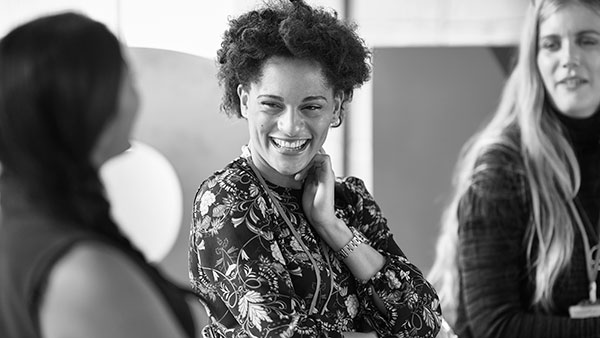 Three people engaging in conversation, smiling at each other in a casual setting. The central person has curly hair and wears a patterned outfit. The image is in black and white, focusing on expressions and interaction.