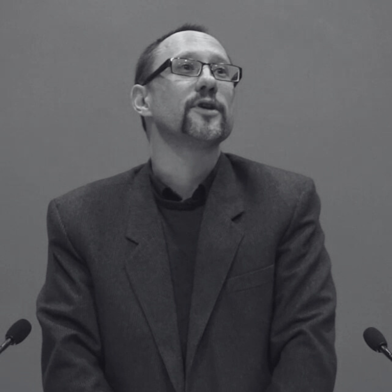 An image of Alan Finlayson with glasses and a goatee stands behind a podium, speaking into two microphones. They are wearing a suit jacket and looking upwards. The image is in black and white.