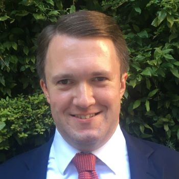 A smiling man in a suit and red tie stands in front of lush green foliage.