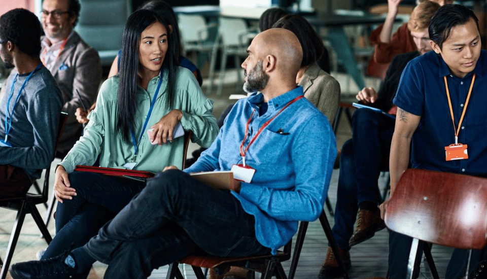 A group of people sitting on chairs in a classroom setting. Two individuals in the foreground are engaged in conversation, while others are focused on different activities. They all have lanyards, suggesting a conference or workshop environment.