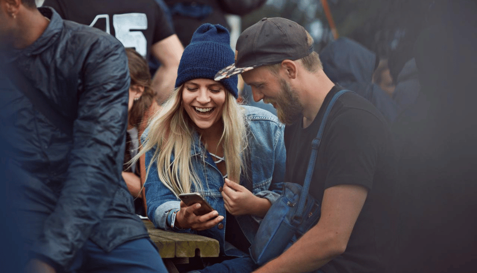 A young woman and man are laughing while looking at a smartphone outdoors. She has long blonde hair and wears a blue beanie and denim jacket. He has a beard and wears a black cap and t-shirt. Other people are blurred in the background.
