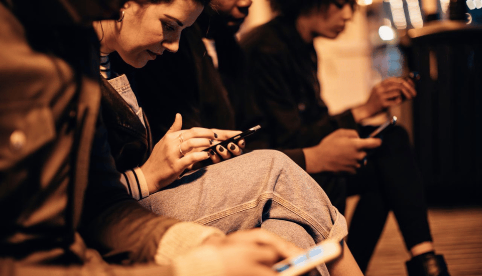 A group of people sitting in a row, focused on their smartphones. The setting appears to be urban and possibly at night, with blurred lights in the background. They are dressed casually and are close together.
