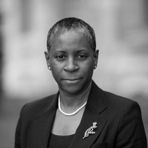 An image of I. Stephanie Boyce with short hair wearing a dark blazer and a pearl necklace, looking directly at the camera with a neutral expression. The background is blurred, highlighting the person as the focal point. The image is in black and white.