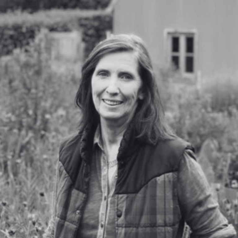 An image of Sue with shoulder-length hair, wearing a vest over a shirt, stands smiling in a garden with a blurred wooden building in the background. The image is in black and white.