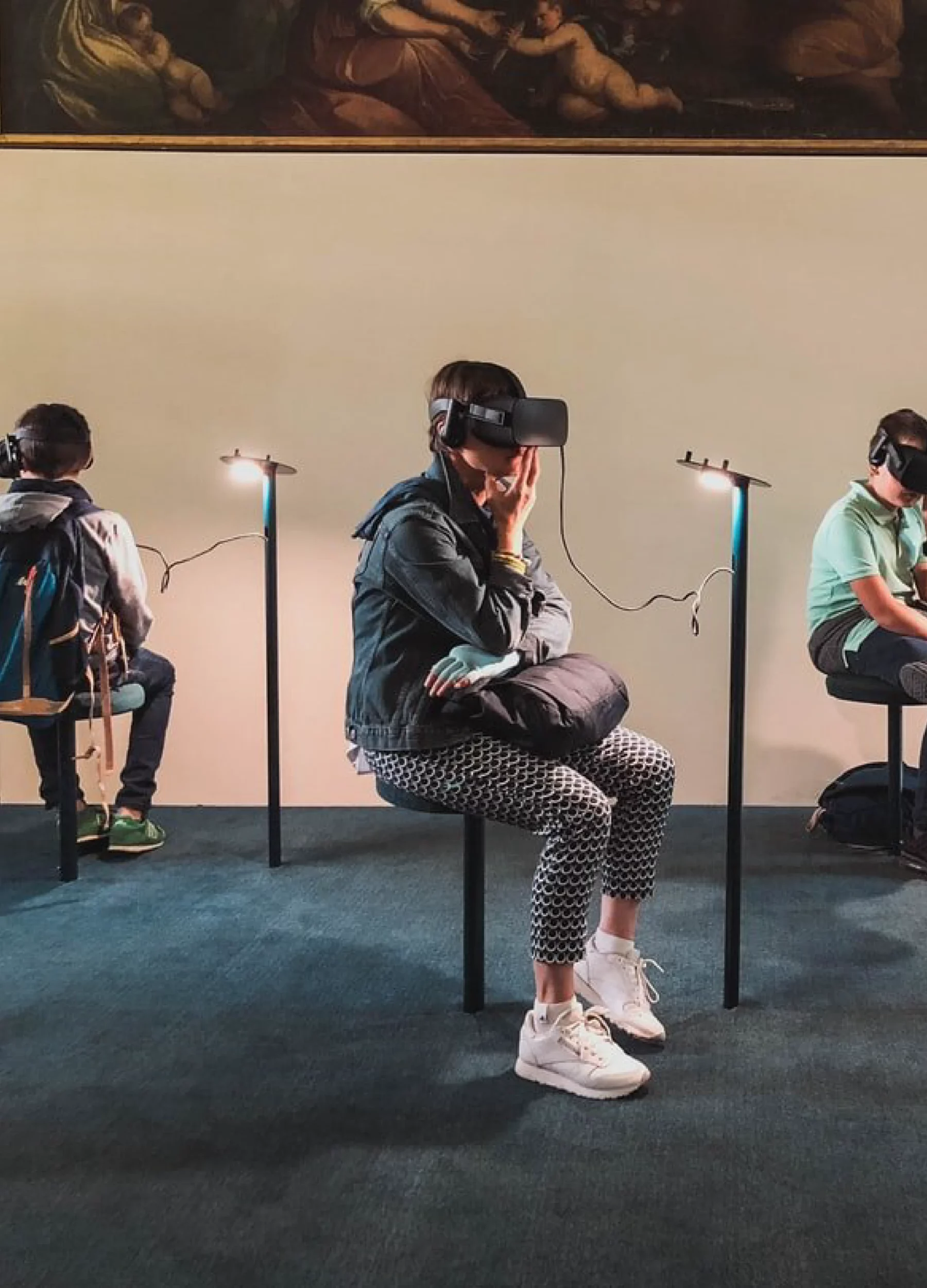Three people sit on stools wearing virtual reality headsets, each connected to a small stand, exploring an innovative digital landscape. In the dimly lit room with a painting above them, the central person sports a denim jacket and patterned pants.