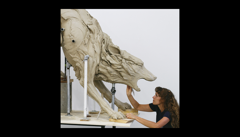 A woman works on a large clay sculpture of a wolf, intricately shaping its fierce, open-mouthed expression. The sculpture is propped up with support rods on a table. The background is plain white.