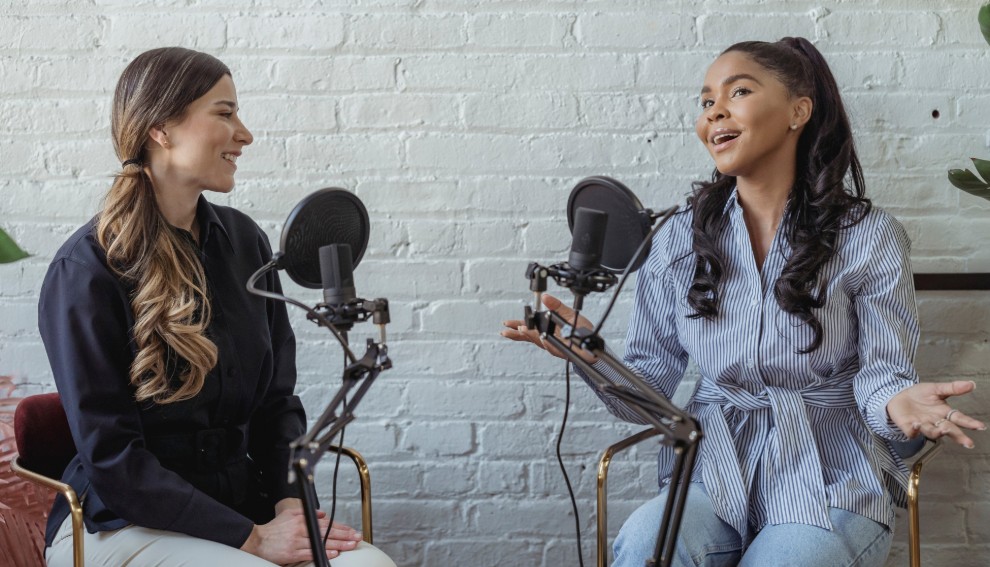 Two women sit facing each other in front of microphones, engaged in a lively conversation about social connections. Both are smiling; one gestures with her hands. They are in a bright room with a white brick wall, suggesting a podcast recording setting.