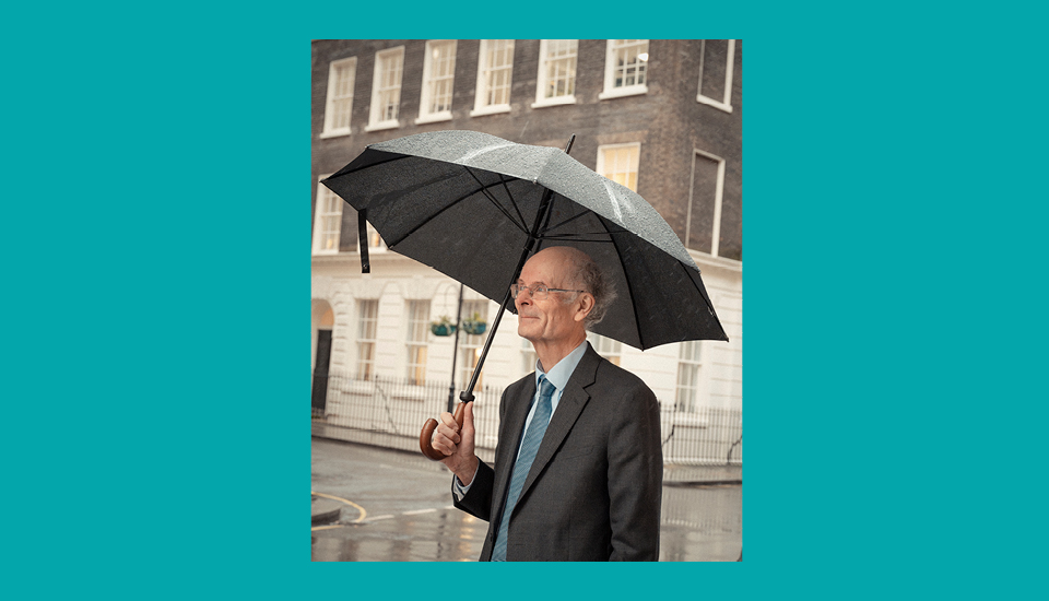 A man in a suit holds an open umbrella, standing on a rainy street in front of a building with large windows. The background is slightly blurred, showing a gray and overcast day.