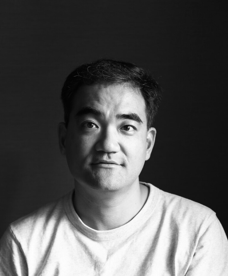 A man with short dark hair wearing a light-colored T-shirt looks slightly upward with a neutral expression. The black-and-white image sets a reflective tone—join us to celebrate the opening of the UK Pavilion at Expo 2025 Osaka.