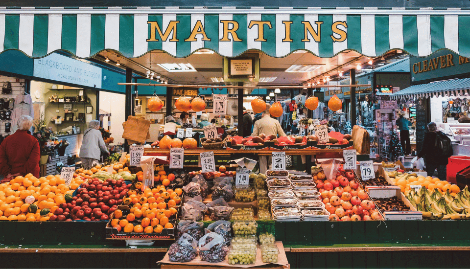 A vibrant market stall under a green and white striped canopy showcases a design for life with its array of colorful fruits and vegetables. The stand, filled with apples, oranges, bananas, and pumpkins, presents clearly marked prices as eager shoppers browse in the background.